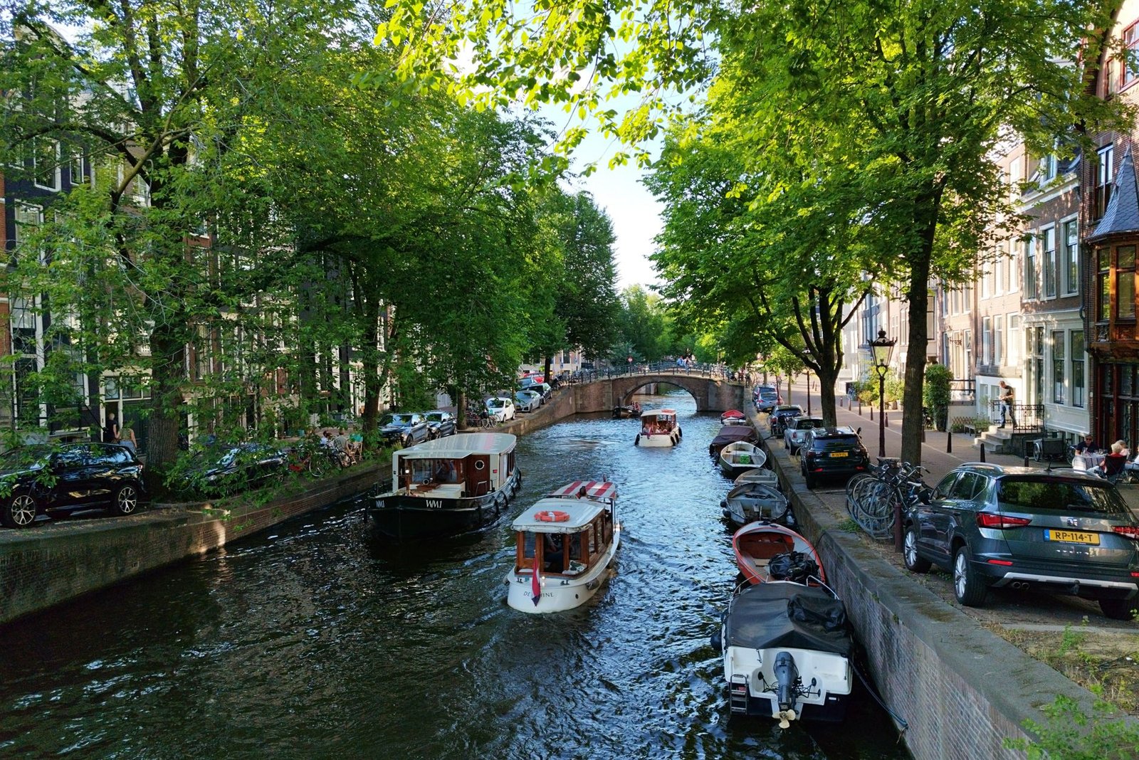 Een levendige zomerdag langs een Amsterdamse gracht, omringd door boten en weelderige bomen.