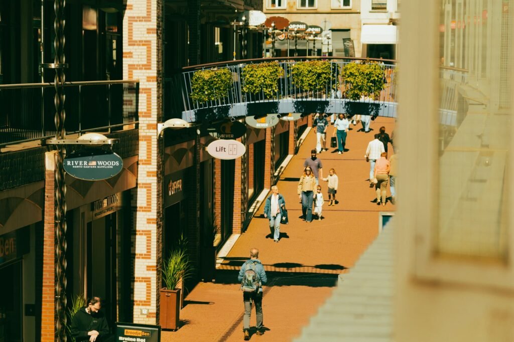 Mensen wandelen door een levendige stadsstraat met bloemen en winkels.